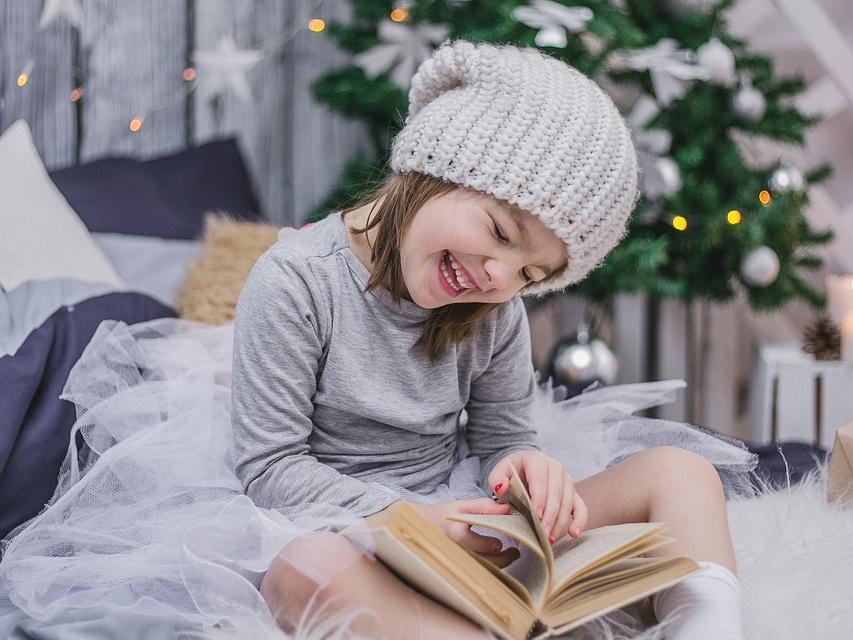 Girl, wearing winter hat, laughing and reading a book in front of  a holiday tree.